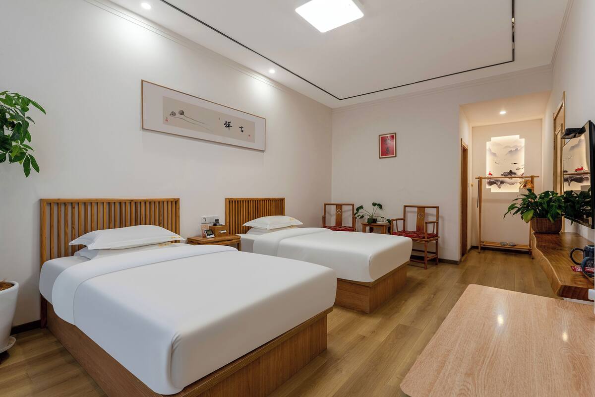 A hotel room with two beds, a wooden headboard, bedside tables, a small desk, a chair, a mirror, and a potted plant.
