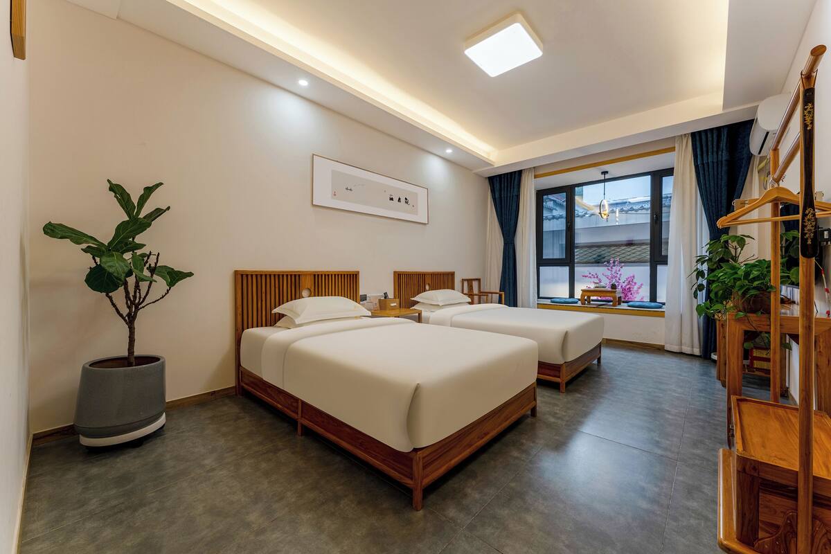 A hotel room with two beds, a potted plant, a bedside table, and a window with curtains.