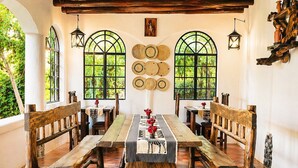 A rustic dining area with wooden benches and a long table, surrounded by arched windows with woven mats on the walls.