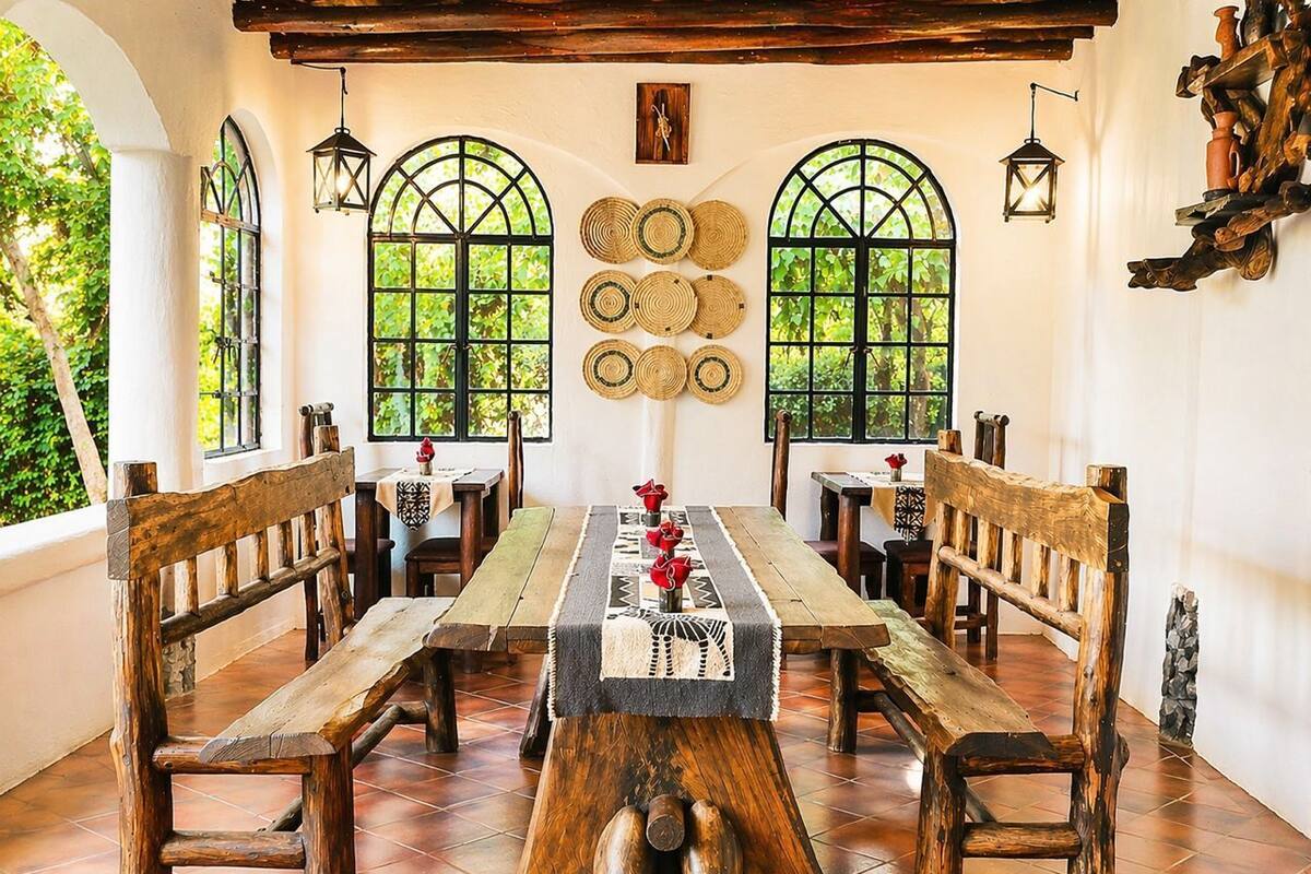 A rustic dining area with wooden benches and a long table, surrounded by arched windows with woven mats on the walls.