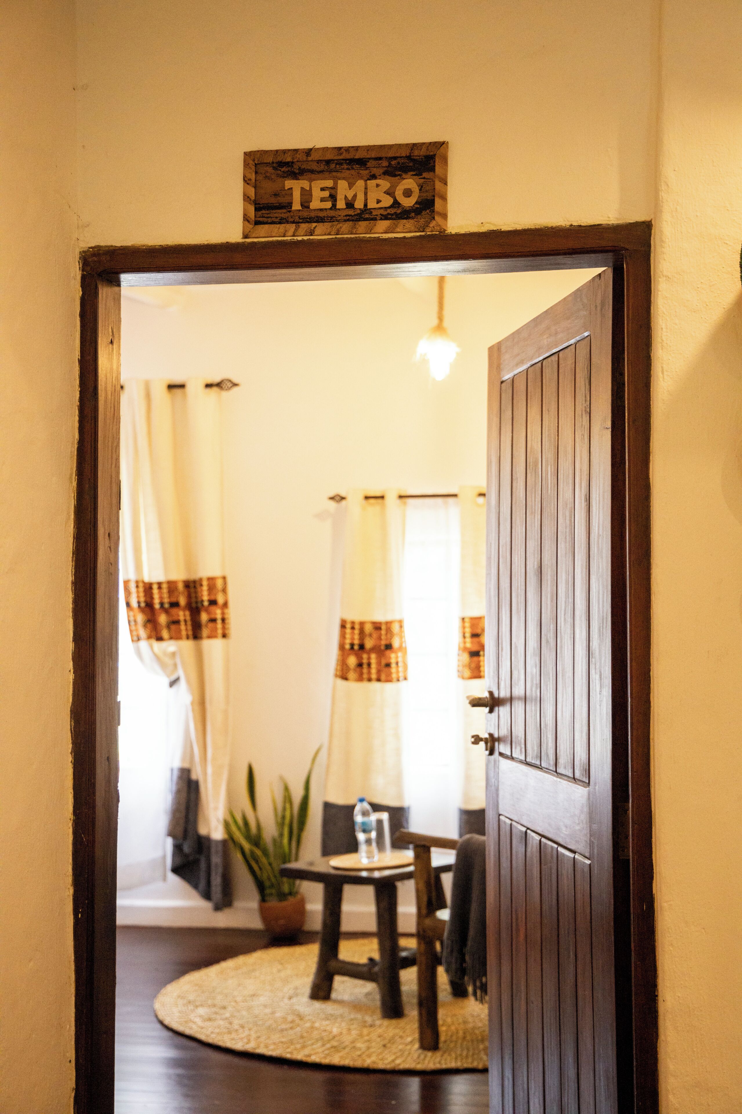 Une porte en bois, sur laquelle est affiché un panneau indiquant « TEMBO », donne accès à une pièce où se trouvent une table ronde, des chaises, une plante en pot et des rideaux.