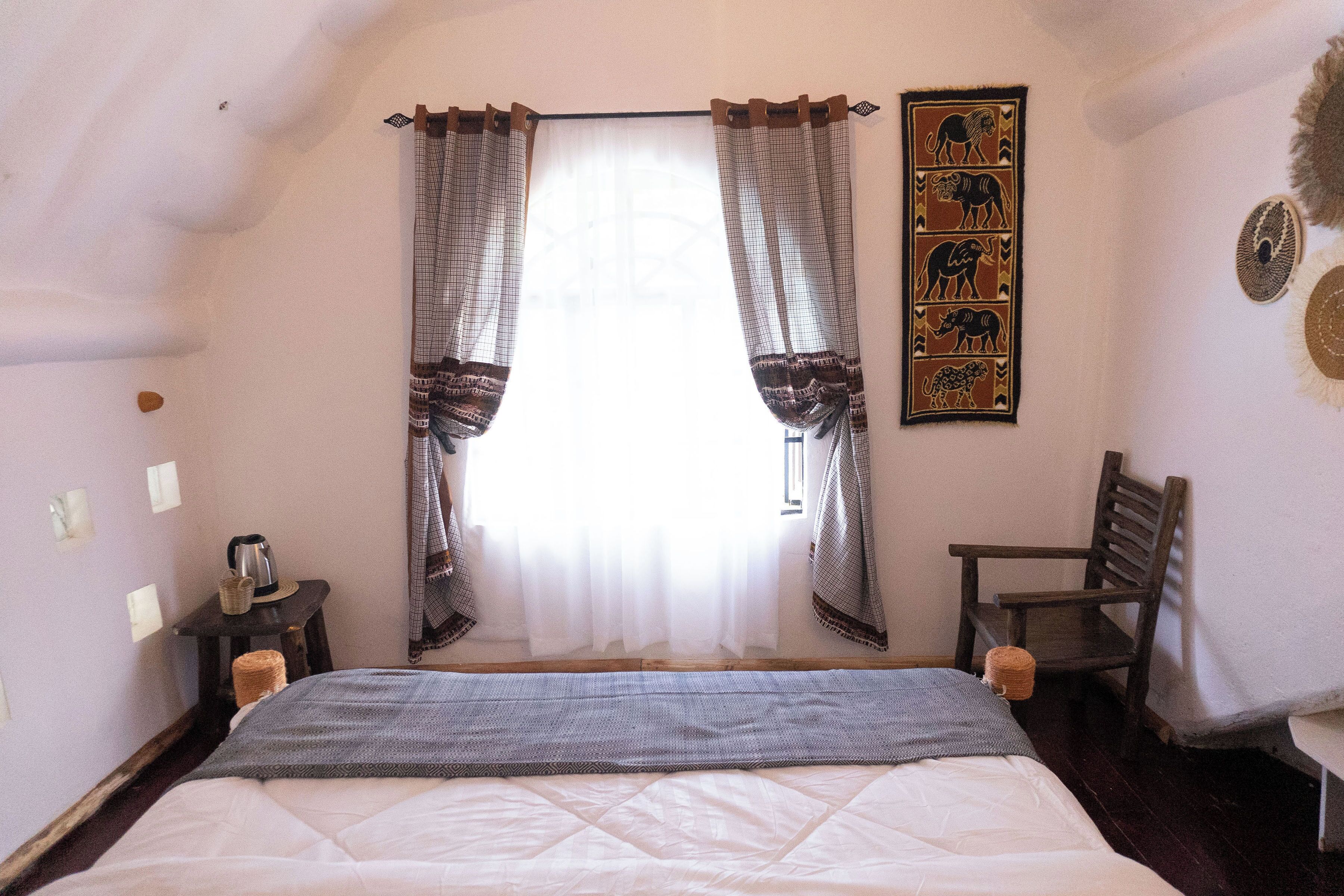A bedroom with a bed, curtains, a chair, a wall art piece, and a small table with a kettle.