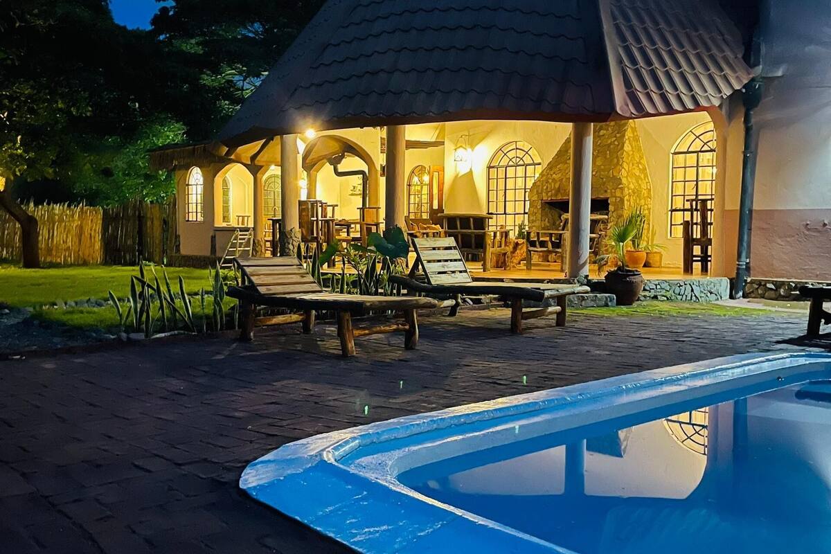 A poolside area with a covered seating area, a stone wall, and a well-lit building with arched windows.