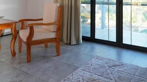 Ein Zimmer mit großem Fenster, Deckenventilator, Holzstuhl, Tisch und einem gemusterten Teppich.