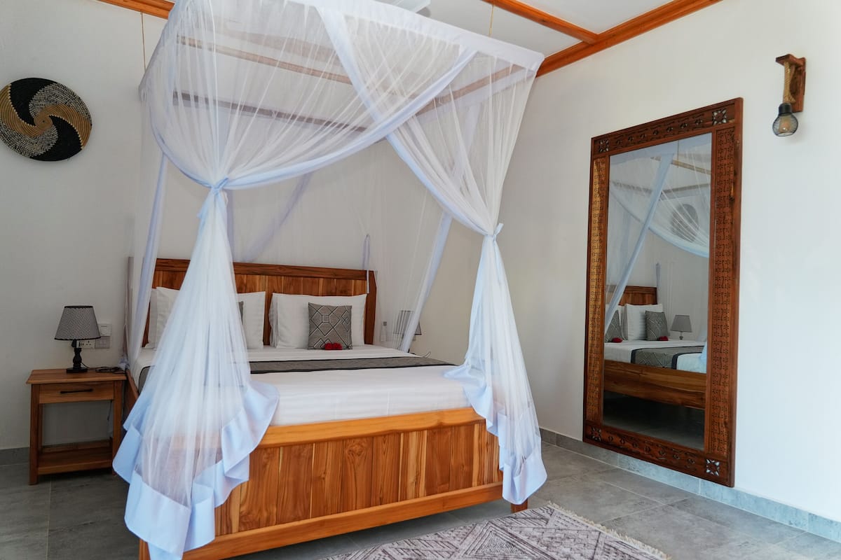 A bedroom with a wooden bed, white bedding, a nightstand with a lamp, a large mirror, and a woven wall hanging.