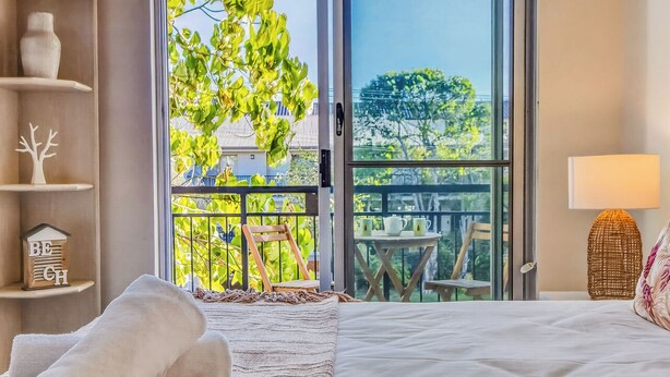 Een slaapkamer met een groot raam, een bed, een nachtkastje, een lamp en uitzicht op een balkon met buitenmeubels.