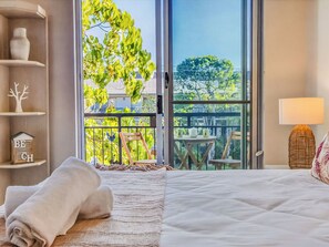 A bedroom with a large window, a bed, a nightstand, a lamp, and a view of a balcony with outdoor furniture. - Coastal Calm at Paradiso - 231 (Kingscliff)