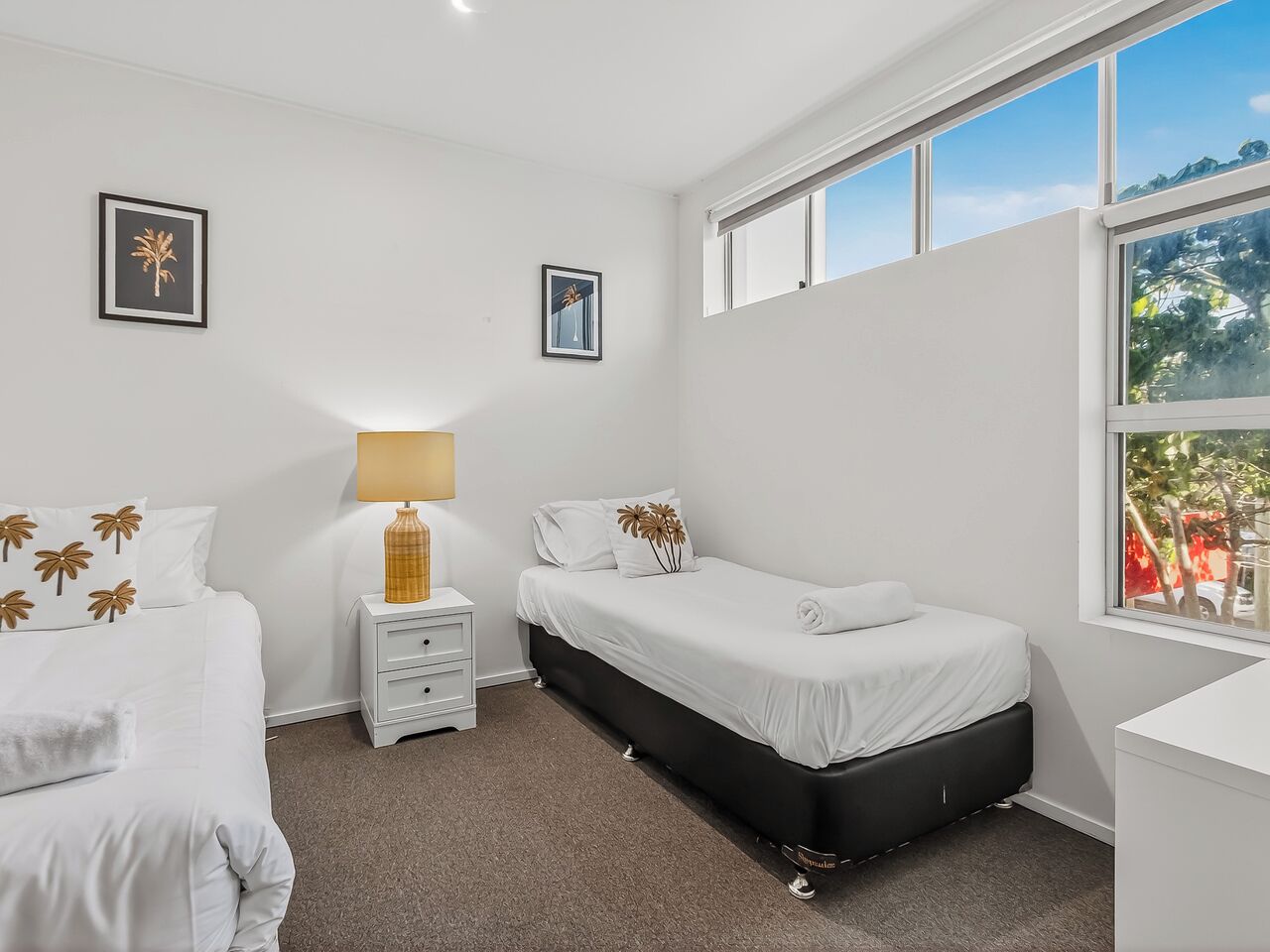 A bedroom with two beds, a bedside table with a lamp, and a window with a view of trees.