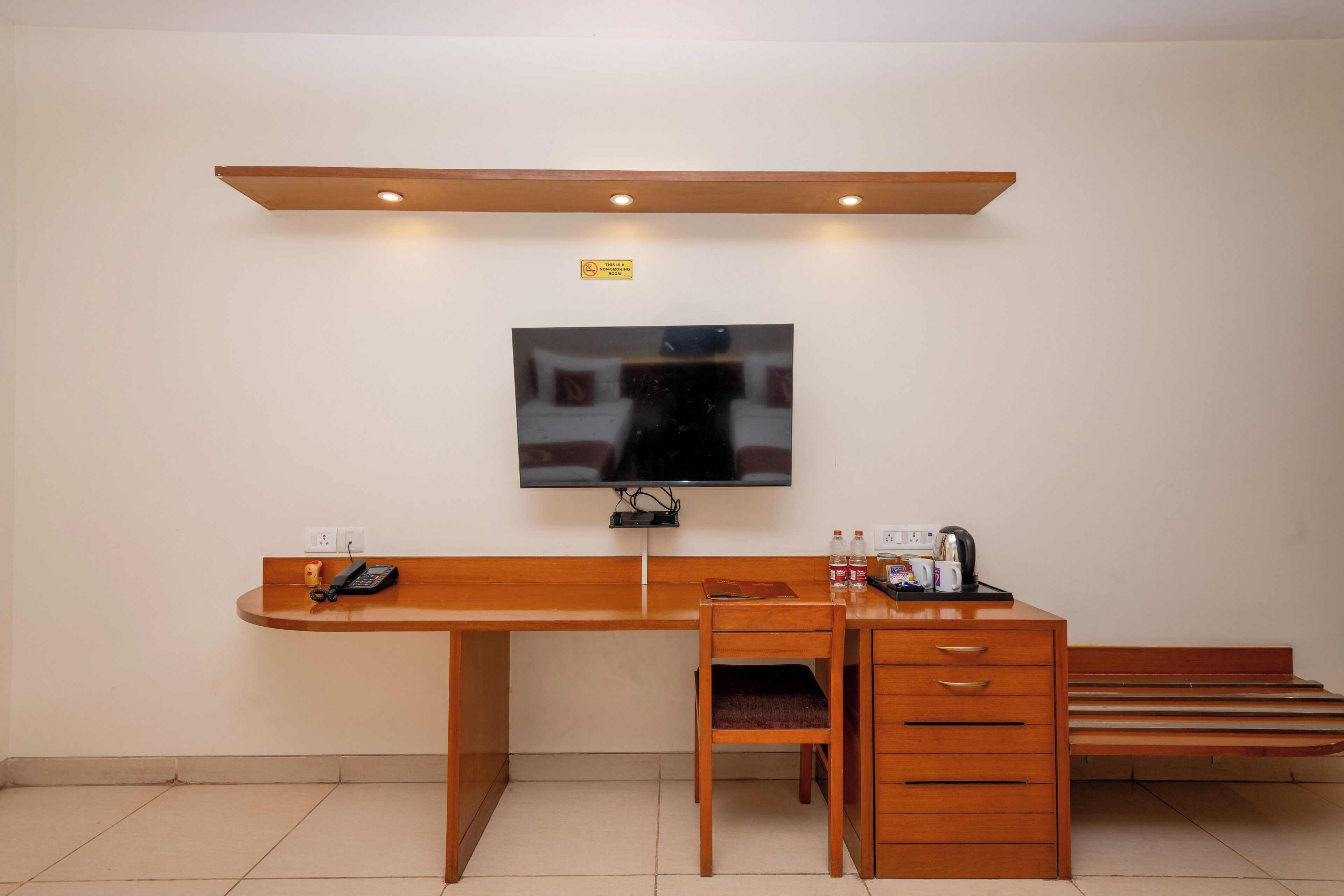 Habitación de hotel con escritorio de madera, una silla, televisor montado en la pared, un mueble bajo con cajones y una cama con cabecera de madera.
