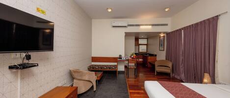 A hotel room with a bed, a desk, a chair, a TV, and an air conditioning unit.