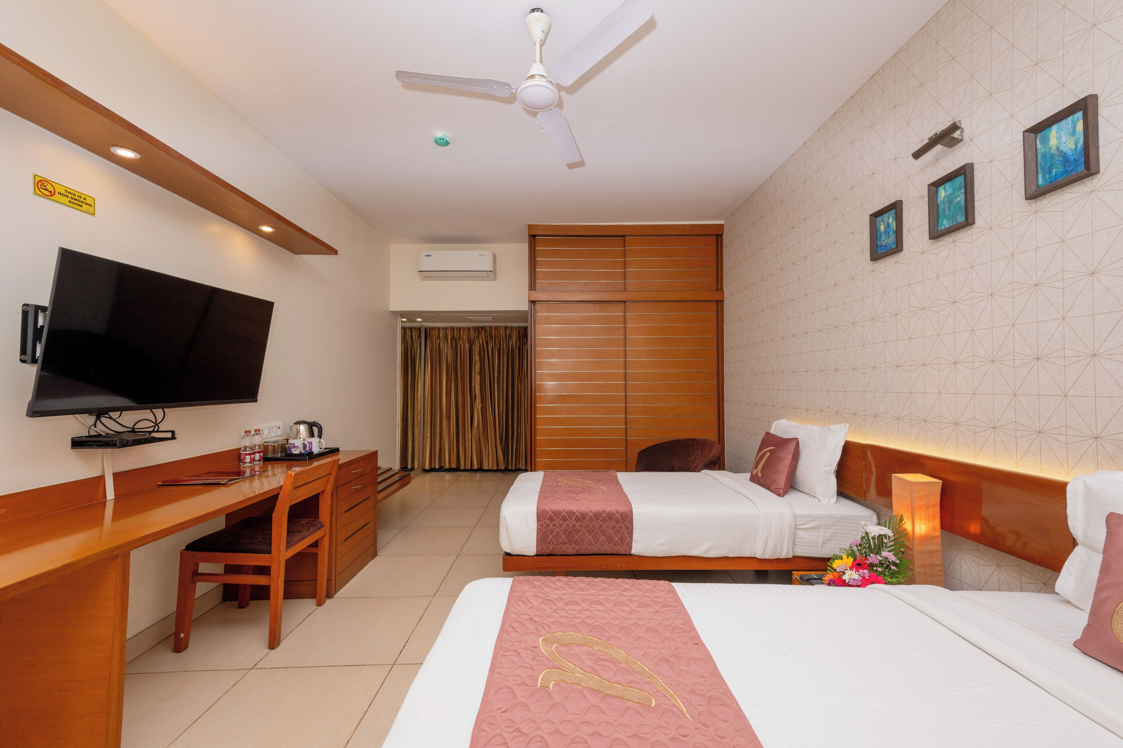 A hotel room with a desk, television, two beds, a ceiling fan, and artwork on the wall.