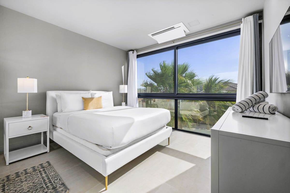 A modern bedroom with a large bed, bedside tables, a desk, and a view of palm trees through a large window.