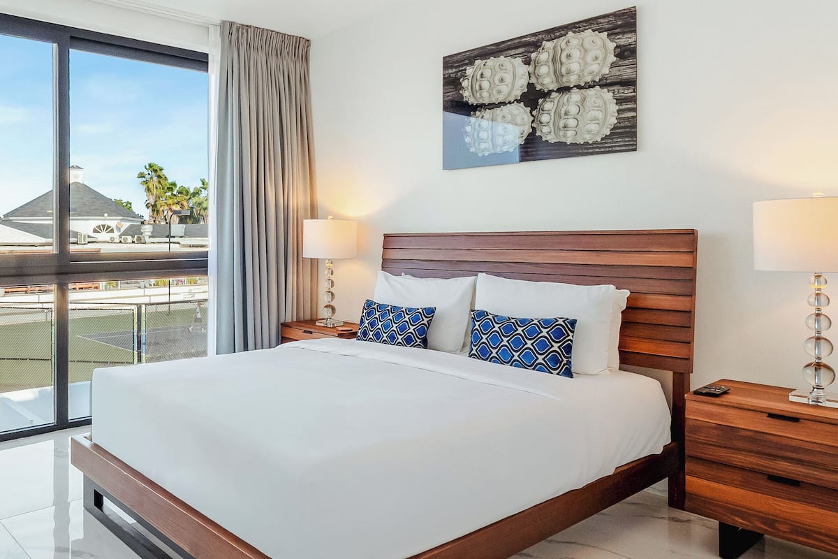 A hotel room with a large bed, wooden headboard, two bedside tables with lamps, a tennis court view, and a decorative wall piece.