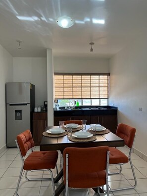 Una cocina moderna con una mesa de comedor para cuatro personas, un refrigerador y una ventana con persianas.