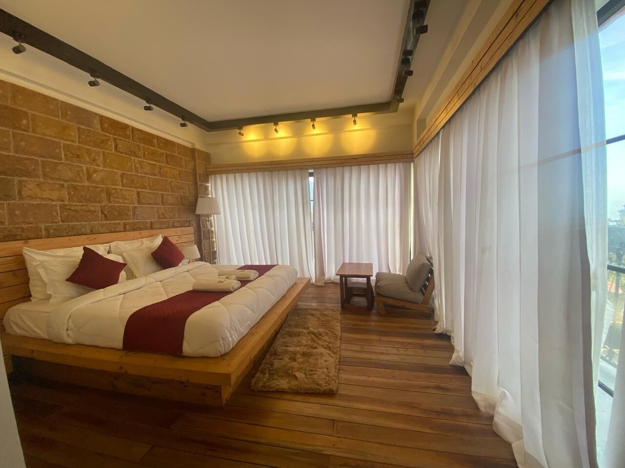 A bedroom with a wooden bed, a small table, a chair, and a view of the city through large windows.