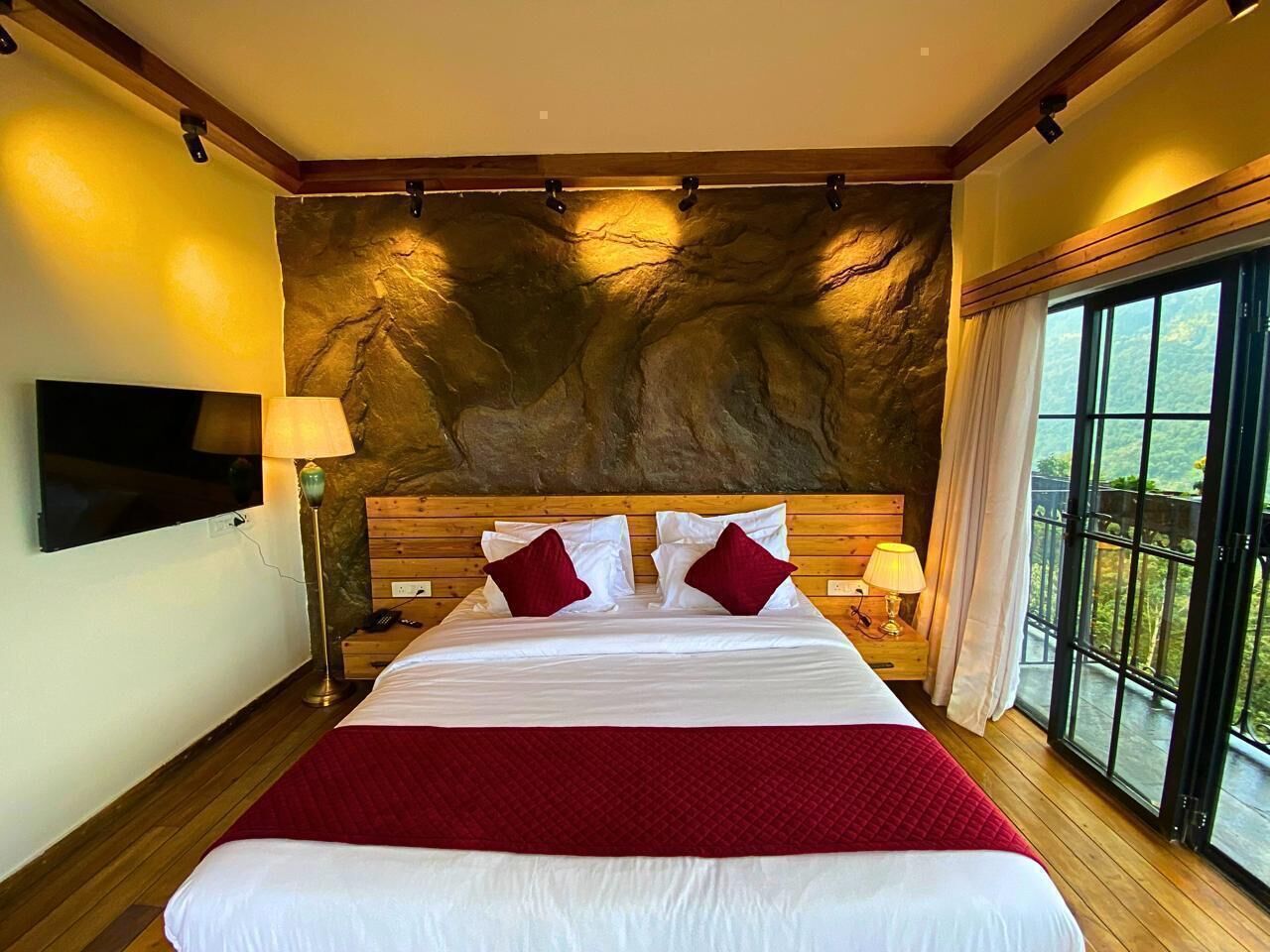 A hotel room with a large bed, wooden headboard, a flat-screen TV, a stone wall, and a balcony with a view.