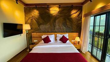 A hotel room with a large bed, wooden headboard, a flat-screen TV, a stone wall, and a balcony with a view.