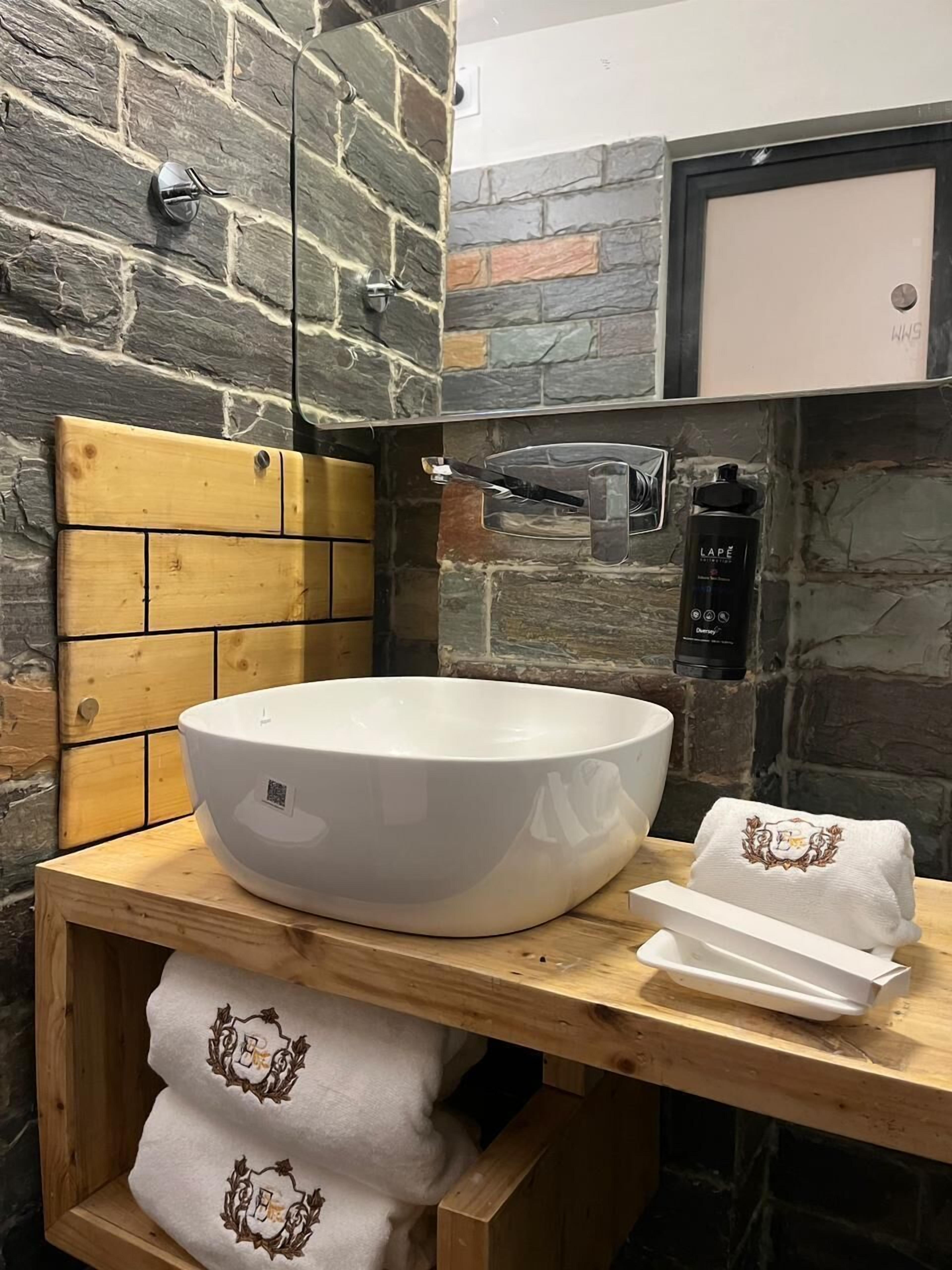 A bathroom with a large white vessel sink, a wooden vanity, a towel rack with towels, and a stone wall.