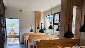 A restaurant with wooden benches and tables, pendant lighting, and a view of a building outside. - Evri Hotels & Resorts (Kolakham)