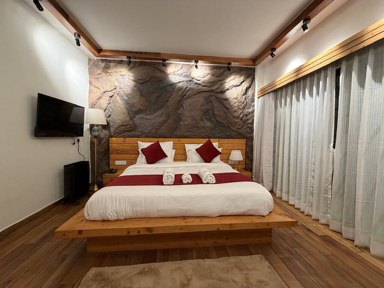Un dormitorio con cama de madera, pared de piedra, televisor de pantalla plana y ventana grande con cortinas.