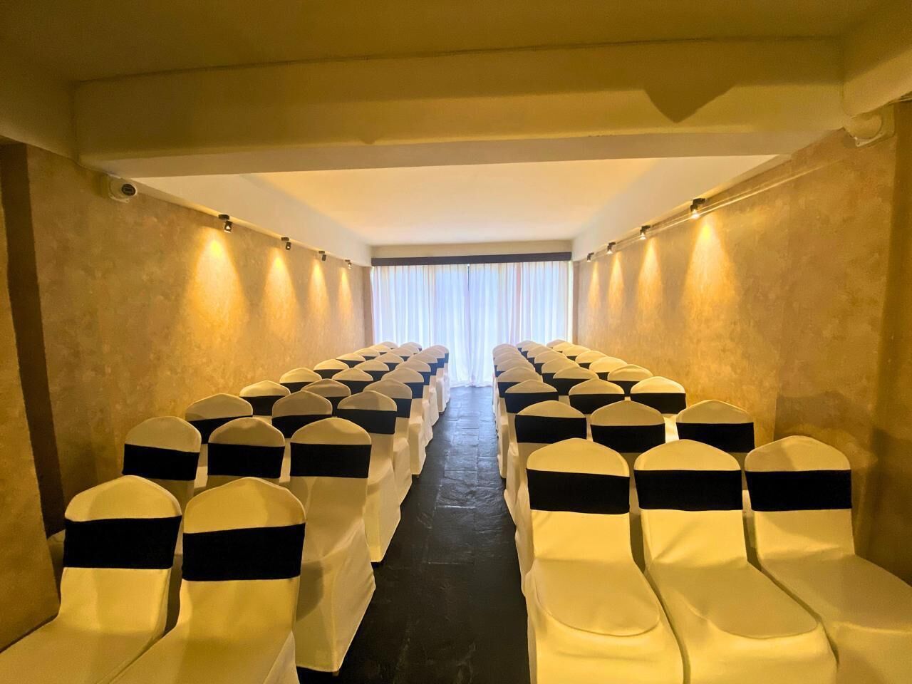 Une rangée de chaises soigneusement disposées, ornées de rubans noirs, un sol sombre et, au fond, une fenêtre avec des rideaux blancs.