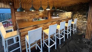 A wooden bar area with a stone wall backdrop, white wicker chairs, and hanging green pendant lights. - Evri Hotels & Resorts (Kolakham)