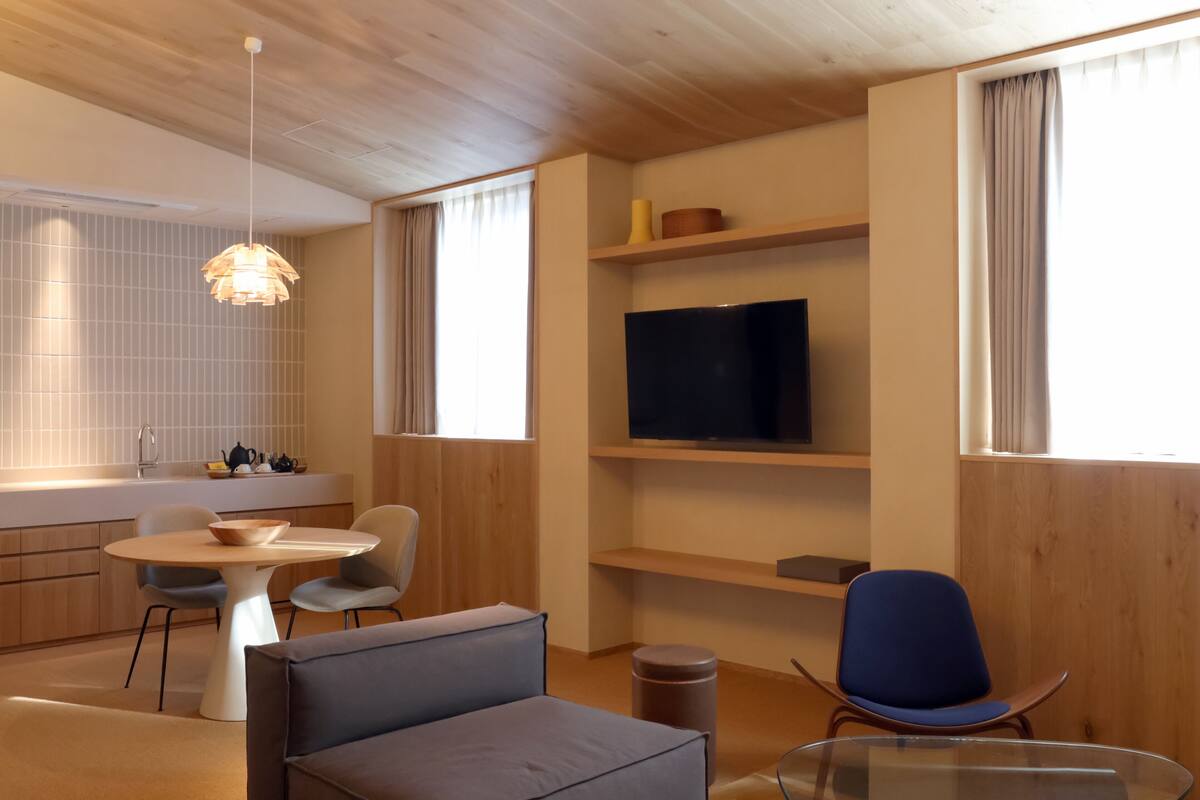 A modern living room with a round table, two chairs, a sofa, a TV on a shelf, and a window with curtains.