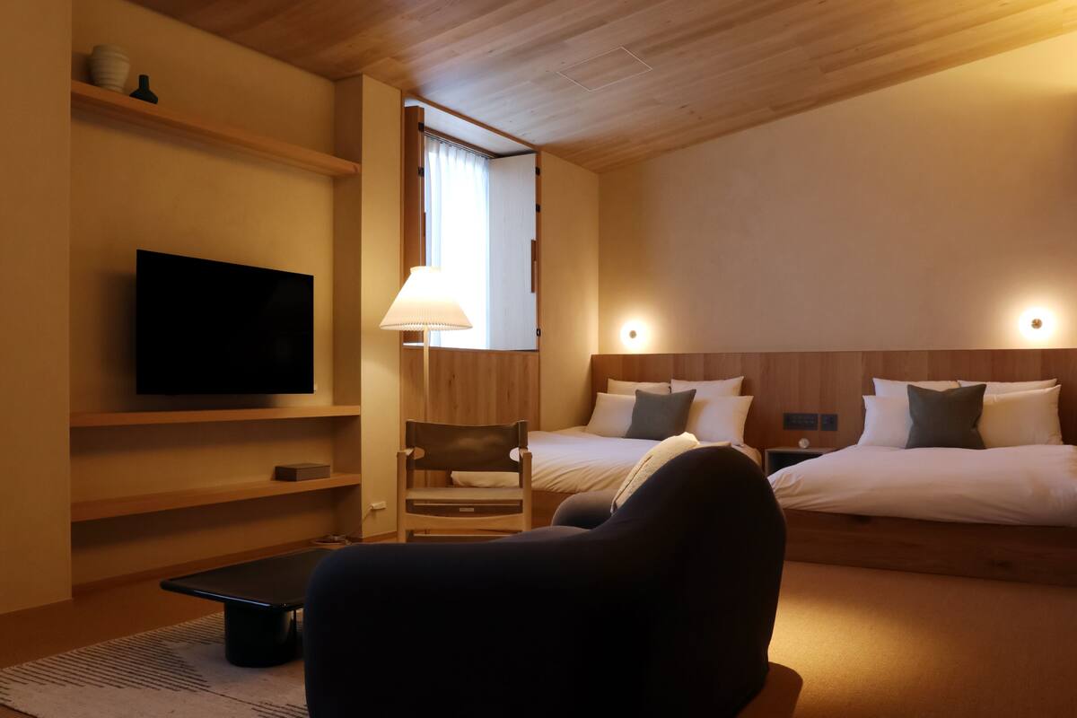A hotel room with a TV, two beds, a chair, a lamp, and a wooden ceiling.