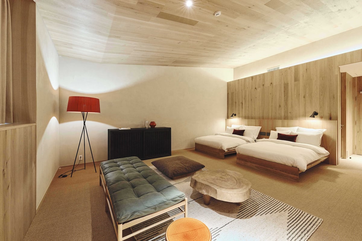 A modern bedroom with a large bed, two smaller beds, a bench, a red floor lamp, and a wooden ceiling.