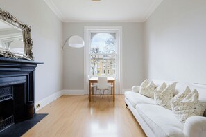 A room with a fireplace, a dining table with chairs, a white sofa with patterned cushions, and a large window with a view of buildings. - Spacious Sun Filled 1BDR Flat in Highbury (Greater London)