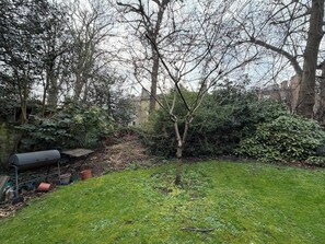 A garden with a variety of trees and shrubs, a barbecue grill, and a small table. - Spacious Sun Filled 1BDR Flat in Highbury (Greater London)