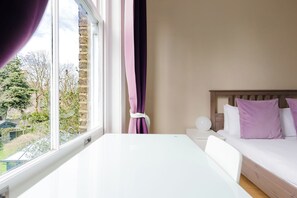 A hotel room with a white table, a bed with white and purple pillows, a window with curtains, and a view of trees and buildings outside. - Spacious Sun Filled 1BDR Flat in Highbury (Greater London)