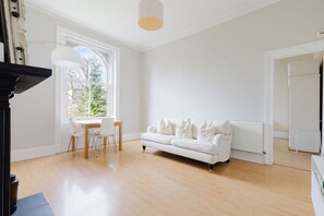 A spacious room with a white sofa, a dining table with chairs, and a large window providing a view of trees. - Spacious Sun Filled 1BDR Flat in Highbury (Greater London)