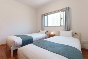 A room with two beds, a window with curtains, a bedside table with a lamp, and a power outlet on the wall. - ASHIBINA INN (Ishigaki)