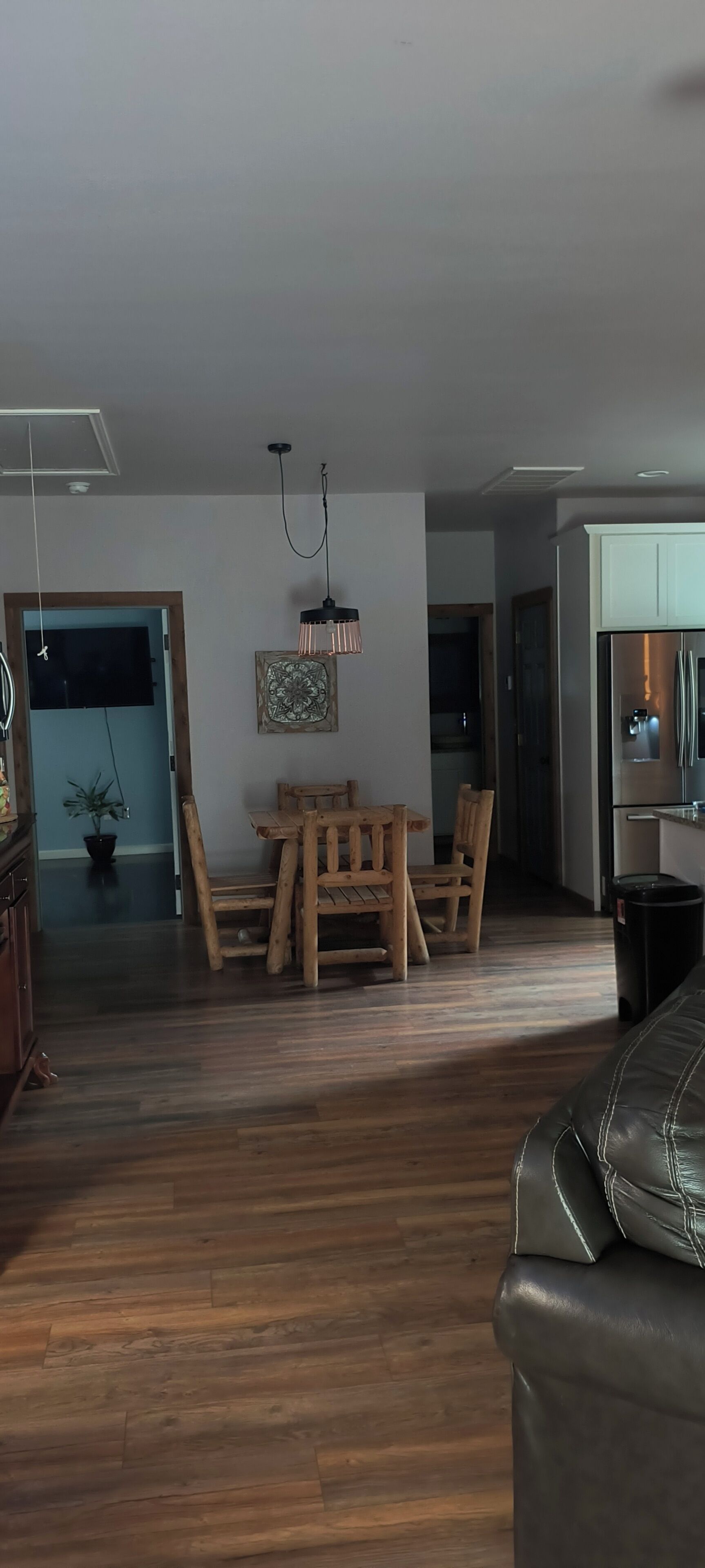 A dining area with a wooden table and chairs, a hanging light fixture, and a kitchen with a refrigerator in the background.