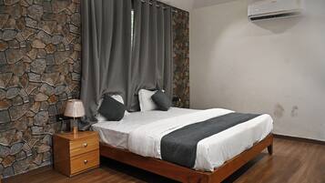 Un dormitorio con pared de piedra, una cama con ropa blanca, cortinas grises, una mesita de noche de madera y un aire acondicionado instalado en la pared.