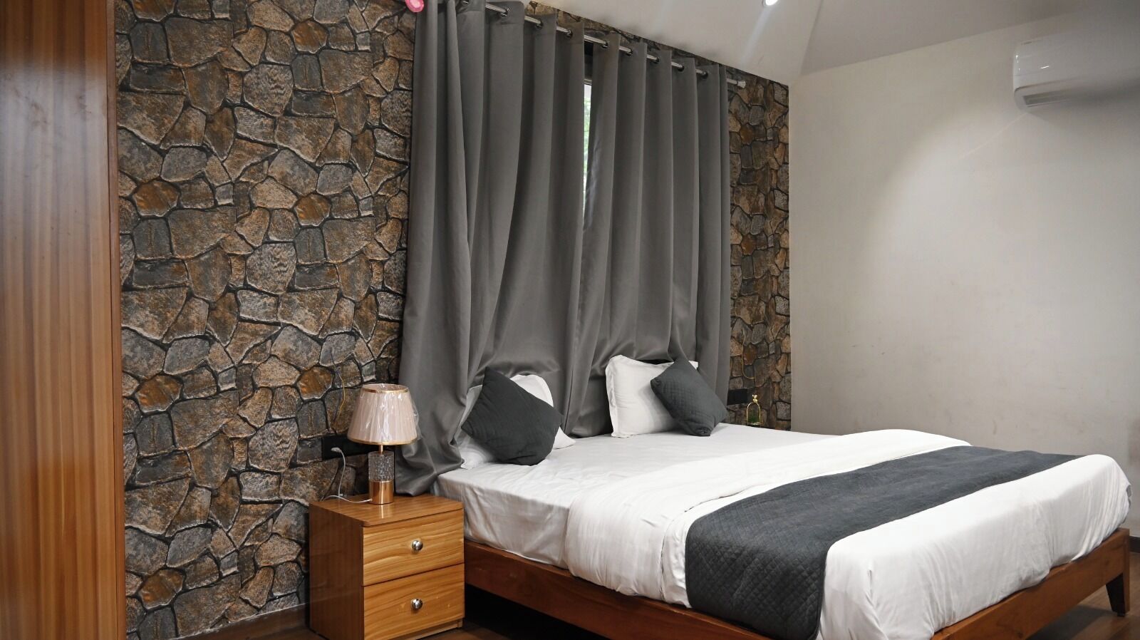 A bedroom with a stone wall, a bed with white bedding, a wooden nightstand, and a lamp.