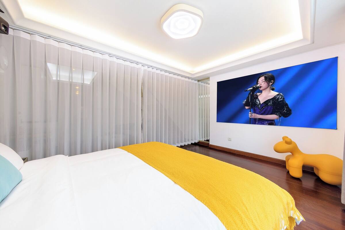 Une chambre d’hôtel avec un lit, un grand écran de télévision, une chaise jaune et un luminaire au plafond.