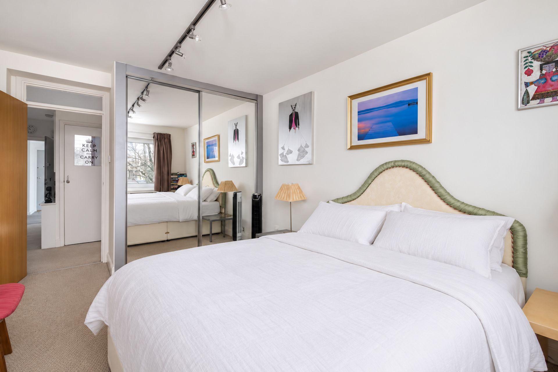 A hotel room with a large bed, a mirror, a bedside table, a lamp, and a painting on the wall.