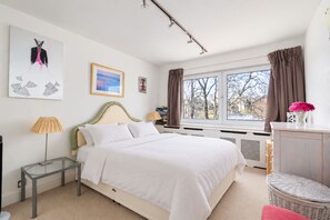 Ein Schlafzimmer mit Bett, Nachttisch, Lampe, Fenster mit Vorhängen und einem Kunstwerk an der Wand.