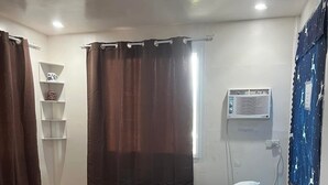 A single bed with a white mattress and pillows, a small shelf unit with decorative items, a window with brown curtains, and a wall-mounted air conditioning unit.