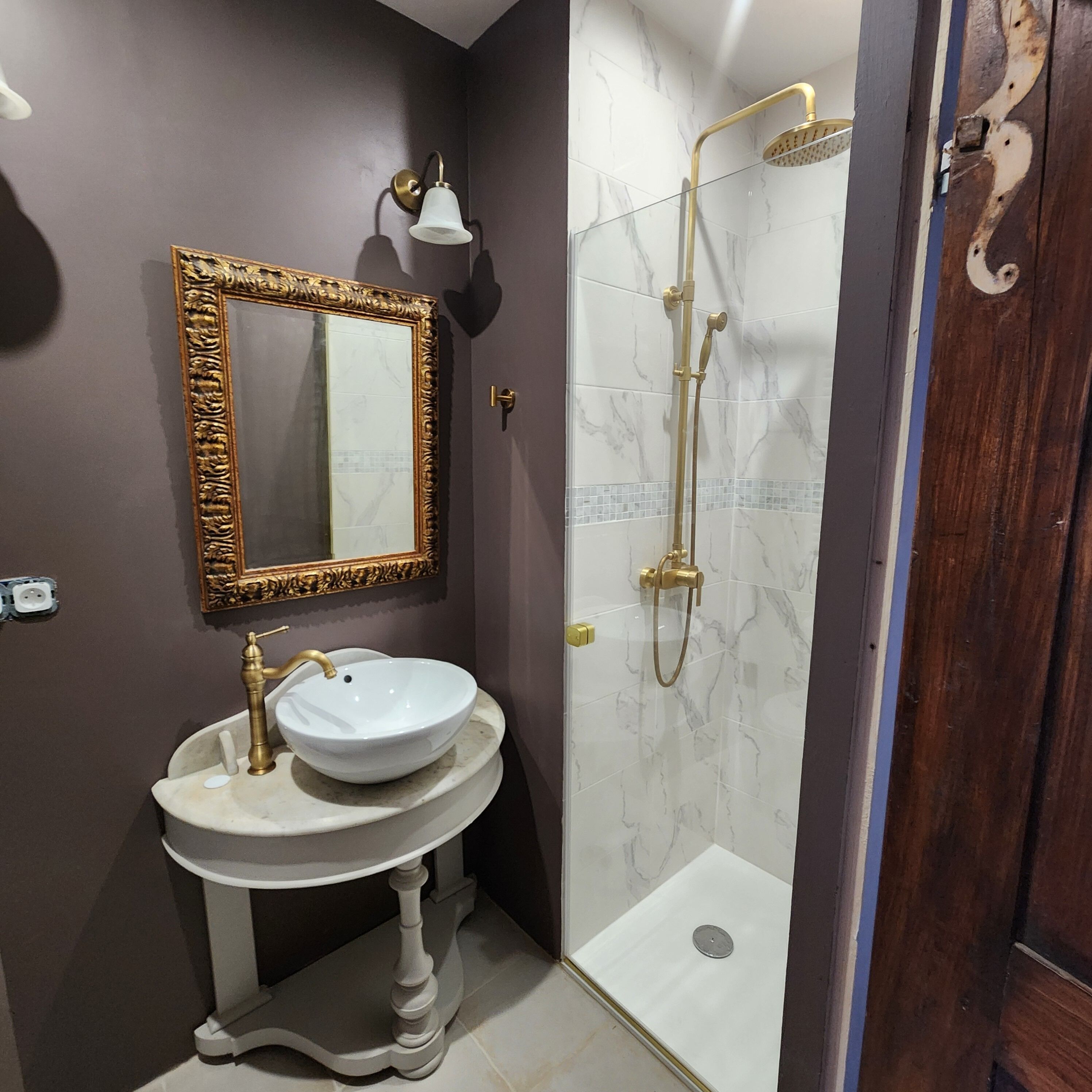 A bathroom with a vanity, a mirror, a shower with a gold fixture, and marble tiles.