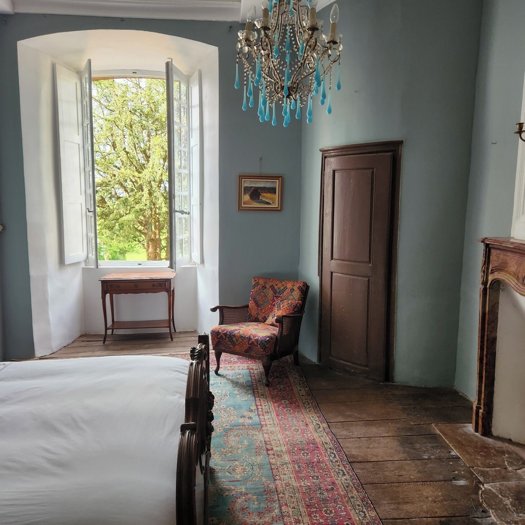 A room with a bed, a chair, a desk, a chandelier, a wardrobe, and a window with a view of trees.