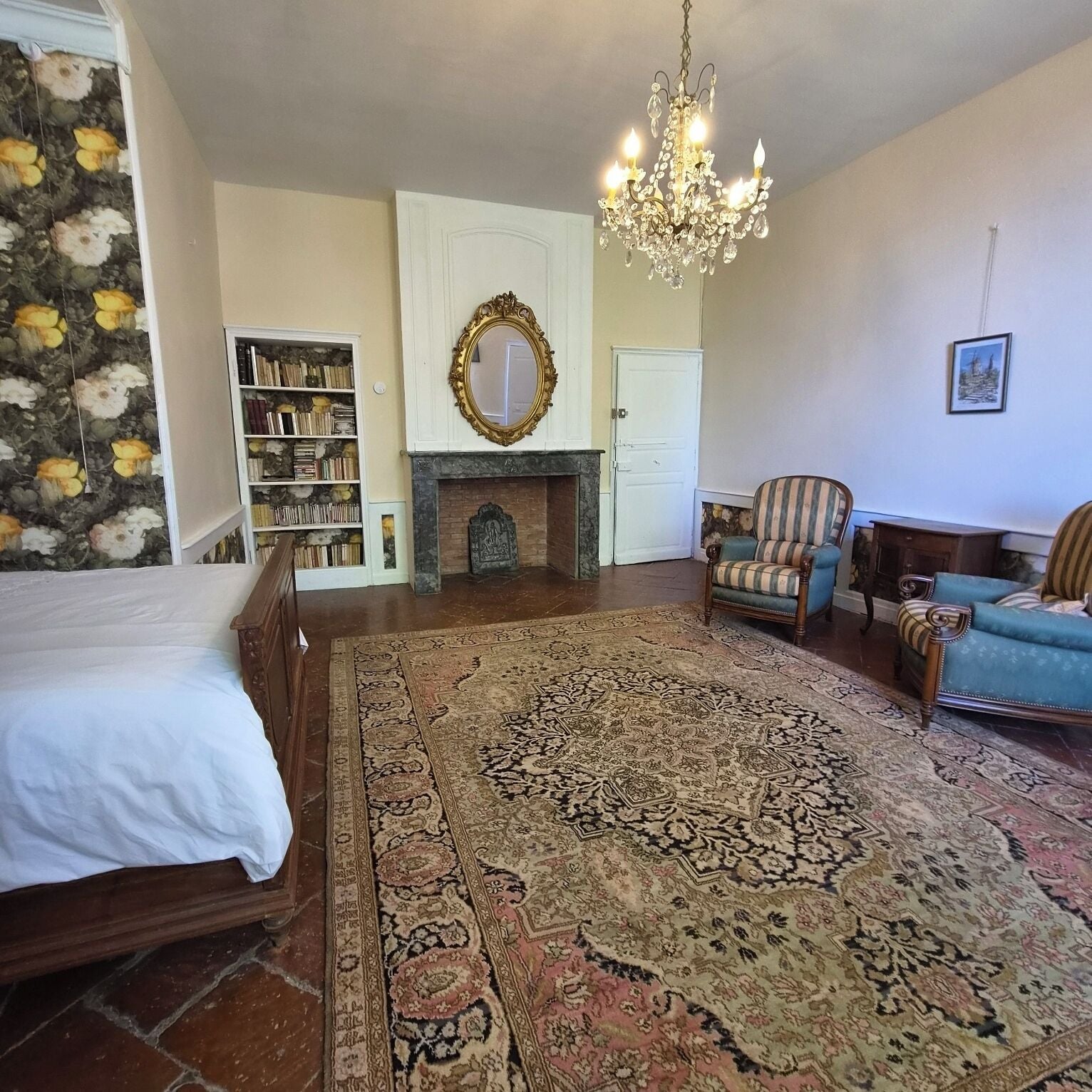 A bedroom with a large bed, a fireplace, a chandelier, a bookshelf, and two armchairs.