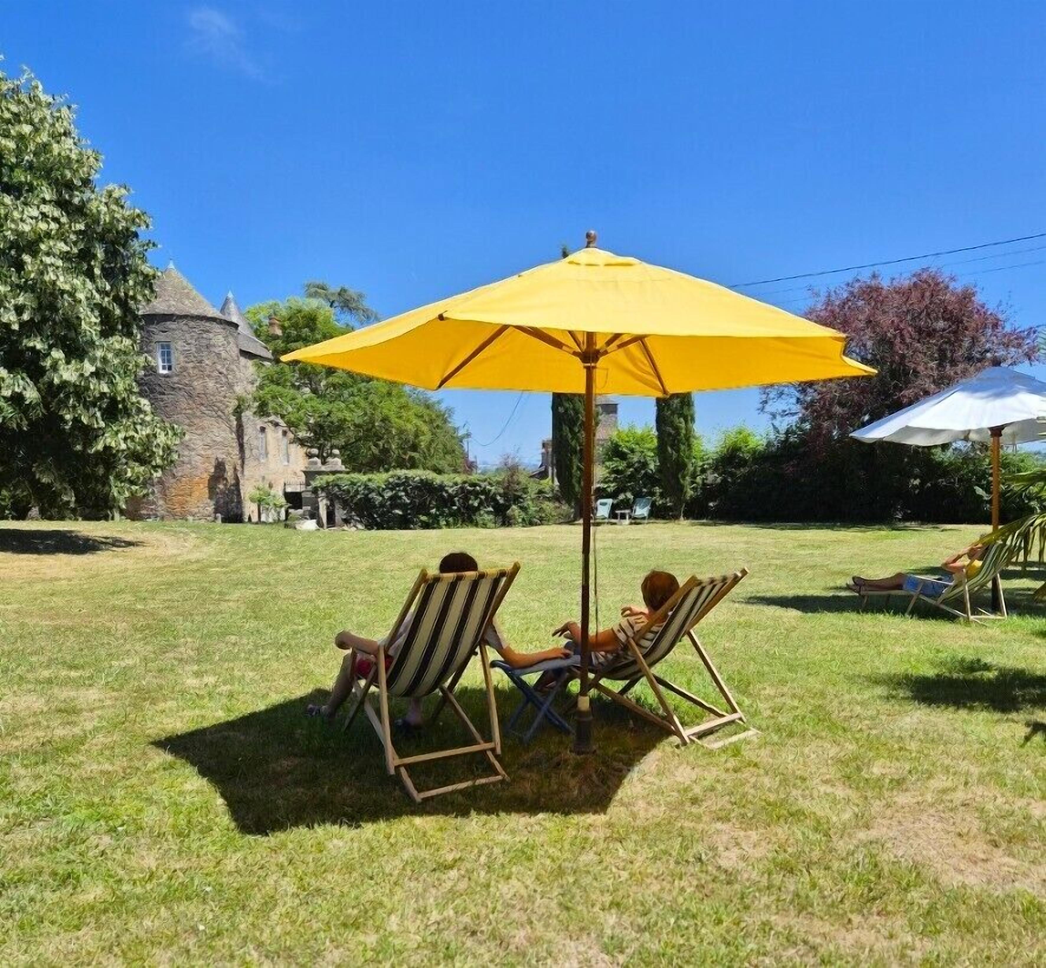 A grassy outdoor area with two lounge chairs under a yellow umbrella, a stone building in the background, and a clear blue sky.