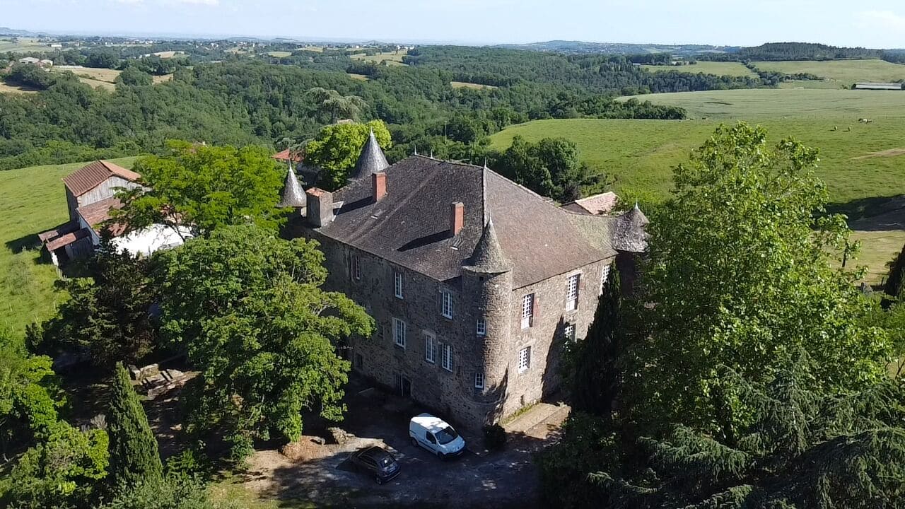 Un grand bâtiment en pierre, doté de nombreuses tours, entouré d’une végétation luxuriante et de champs.