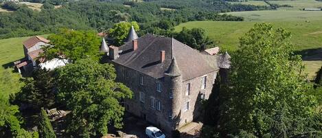 Un grand bâtiment en pierre, doté de nombreuses tours, entouré d’une végétation luxuriante et de champs.