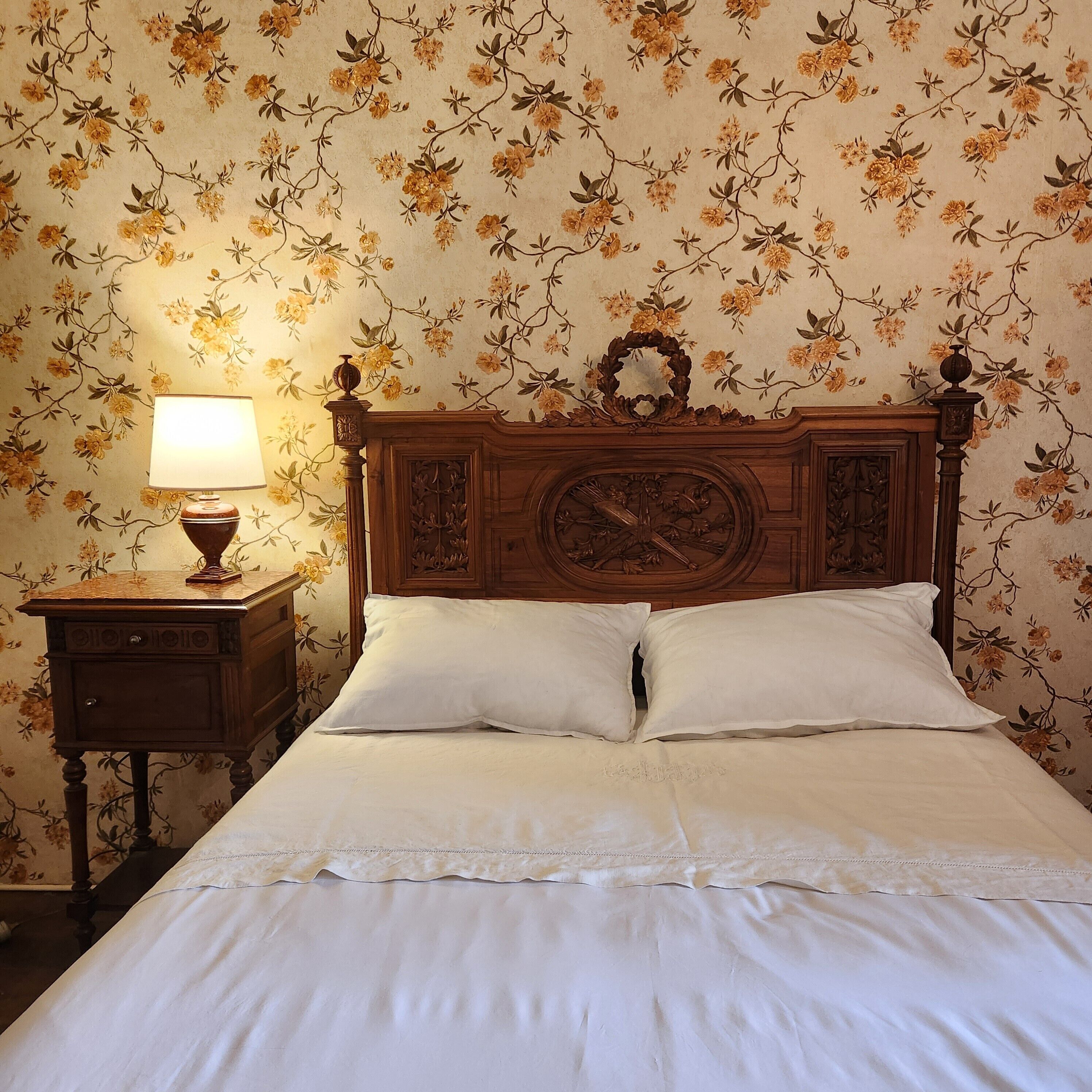 A bedroom with a wooden headboard, a bedside table with a lamp, and floral wallpaper.