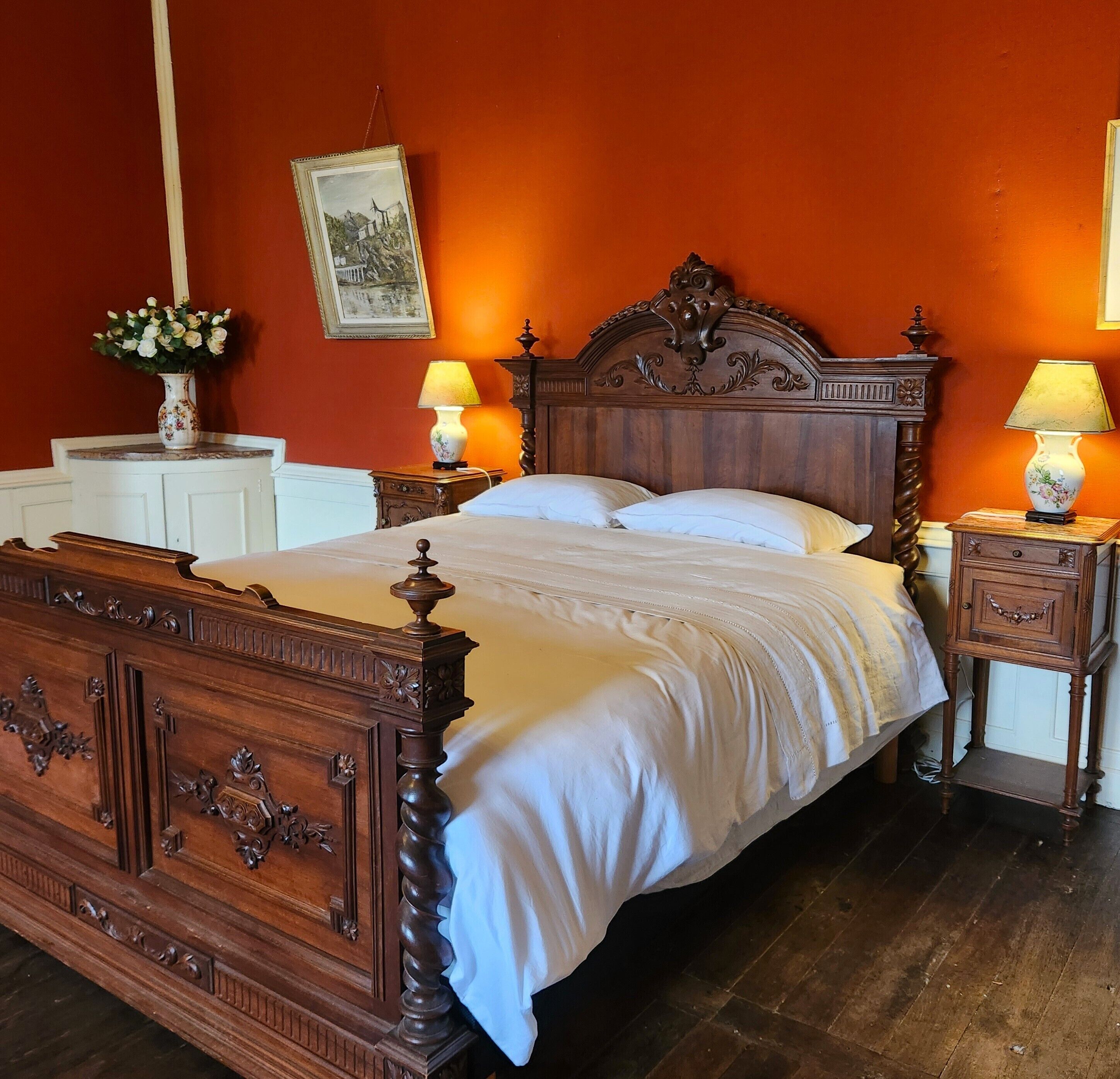 A wooden bed with a headboard, two bedside tables with lamps, a nightstand, and a vase with flowers.