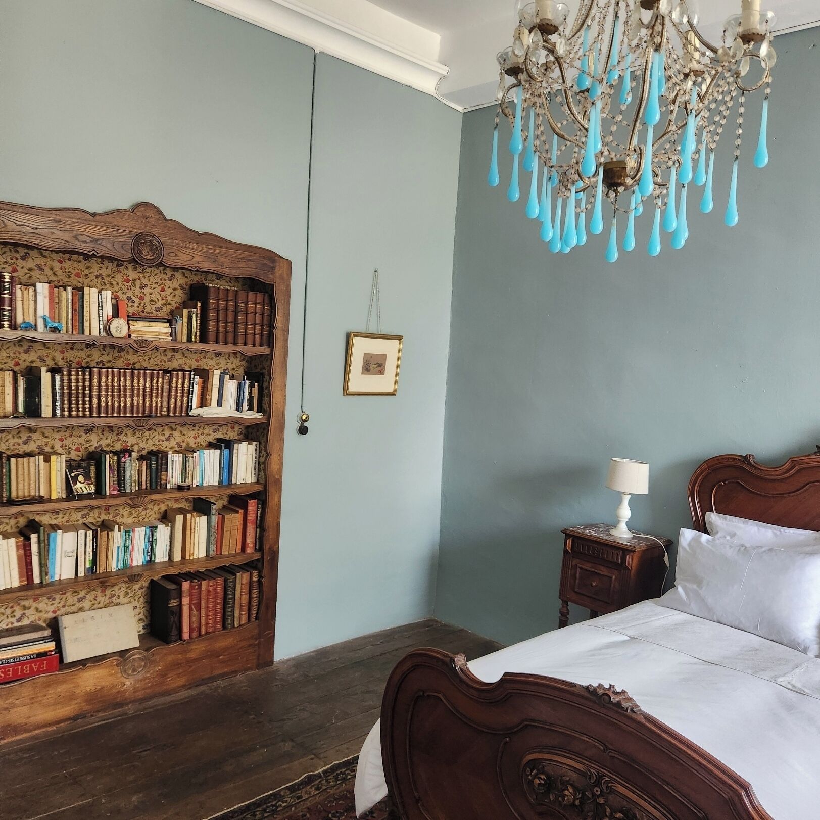A bedroom with a large wooden bookshelf, a chandelier, a bedside table with a lamp, and a bed with white linens.